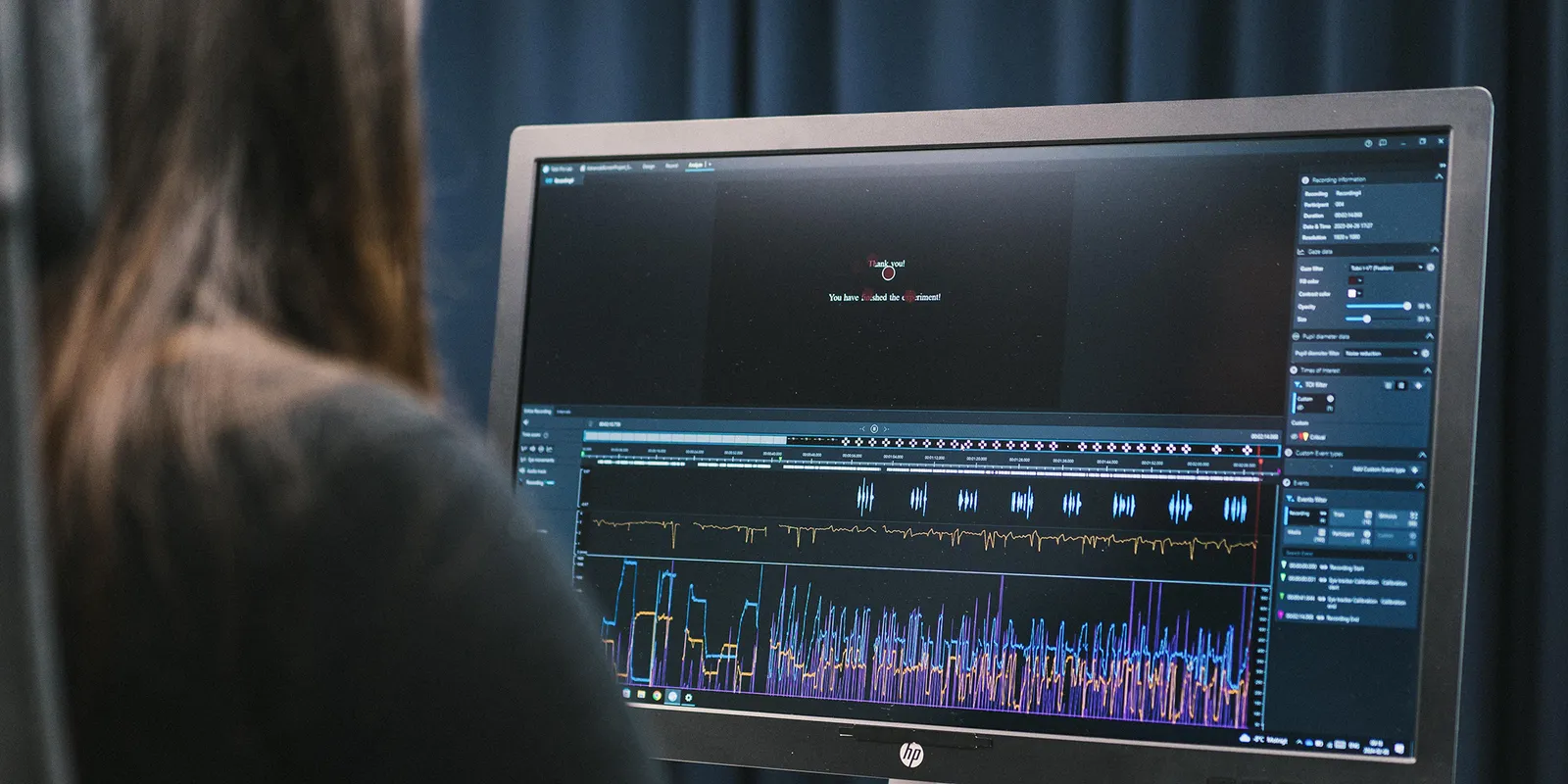 Badaczka przed monitorem z Tobii Pro Lab — na ekranie przebiegi sygnałów zapisanych podczas badania