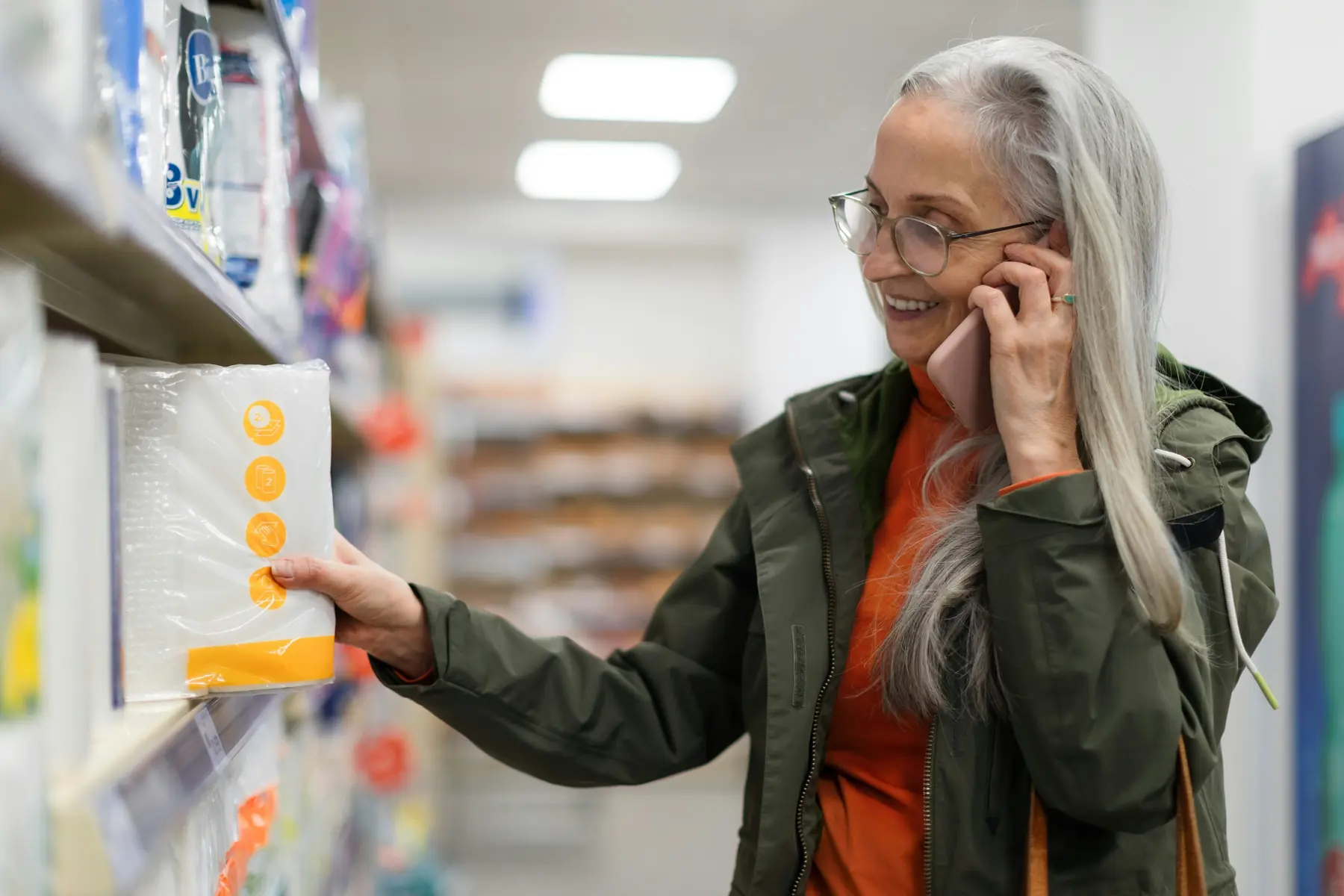 A shopper in a store reaches for a product on a shelf while talking on the phone
